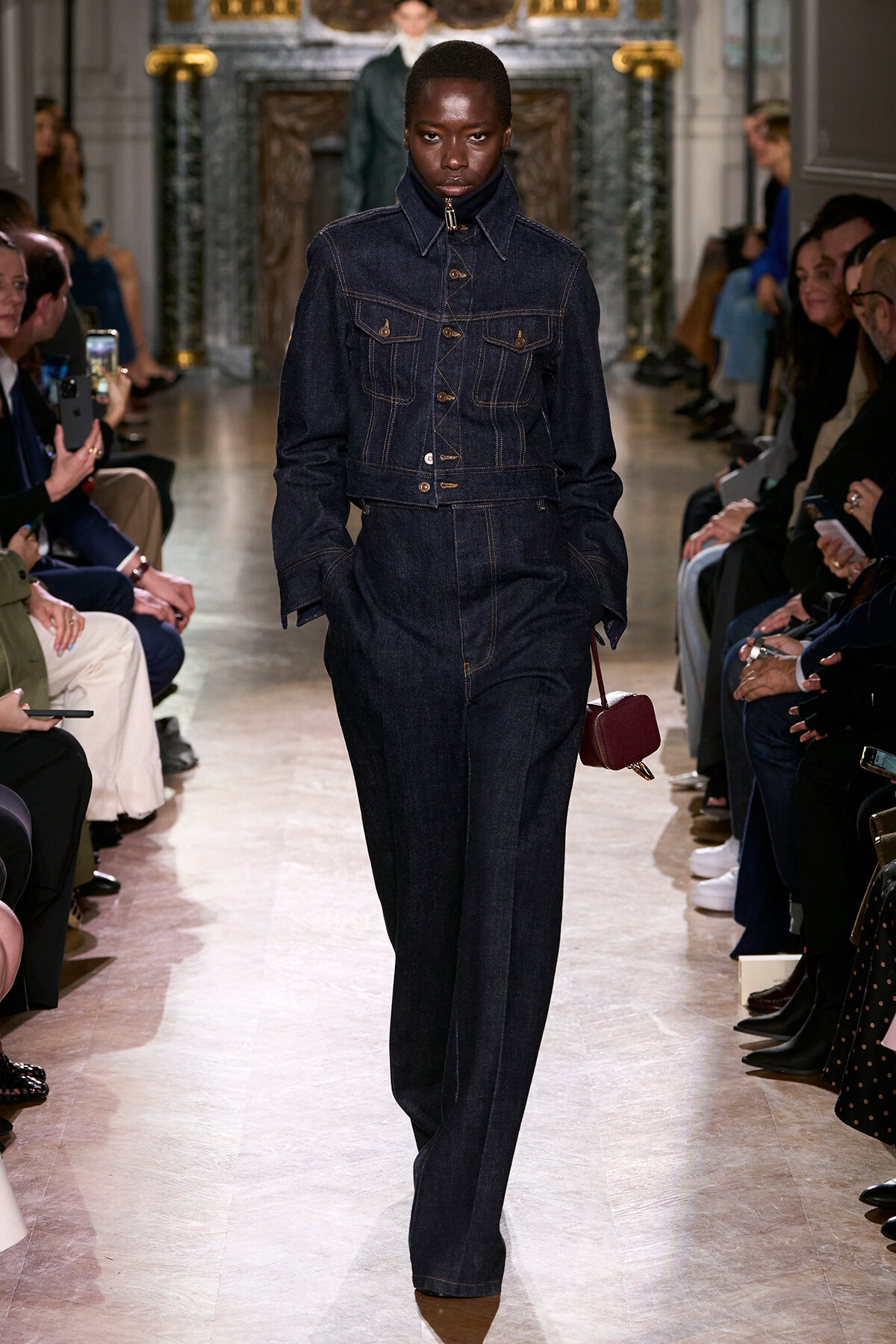 Model in a dark denim jacket and jeans walks the runway at a fashion show, carrying a small burgundy purse.