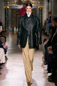 Model walks the runway in a black leather toggle-coat over a white turtleneck and beige wide-leg trousers, with audience on either side.