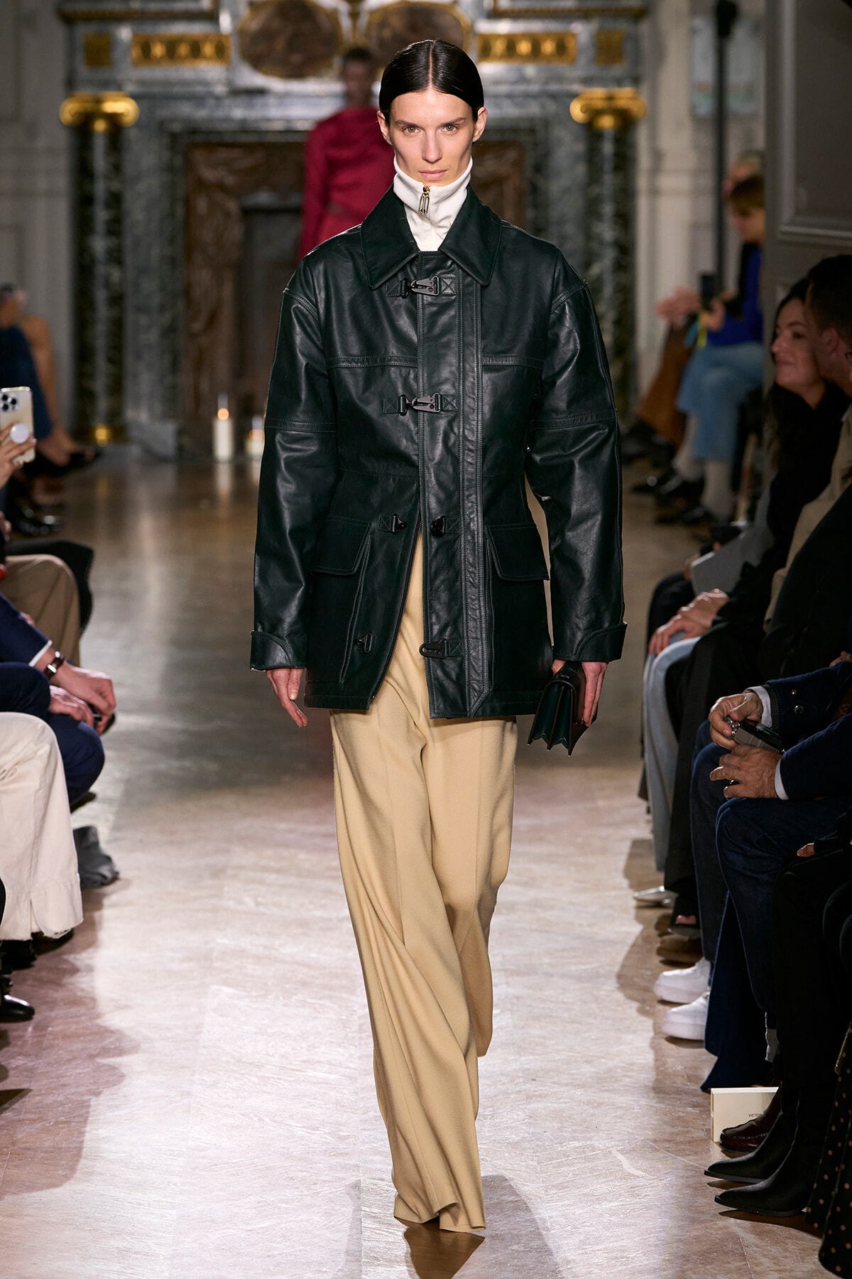 Model walks the runway in a black leather toggle-coat over a white turtleneck and beige wide-leg trousers, with audience on either side.