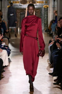 Model walks the runway in a burgundy satin dress with a draped wrap, carrying a small burgundy clutch. Audience lines the catwalk behind.