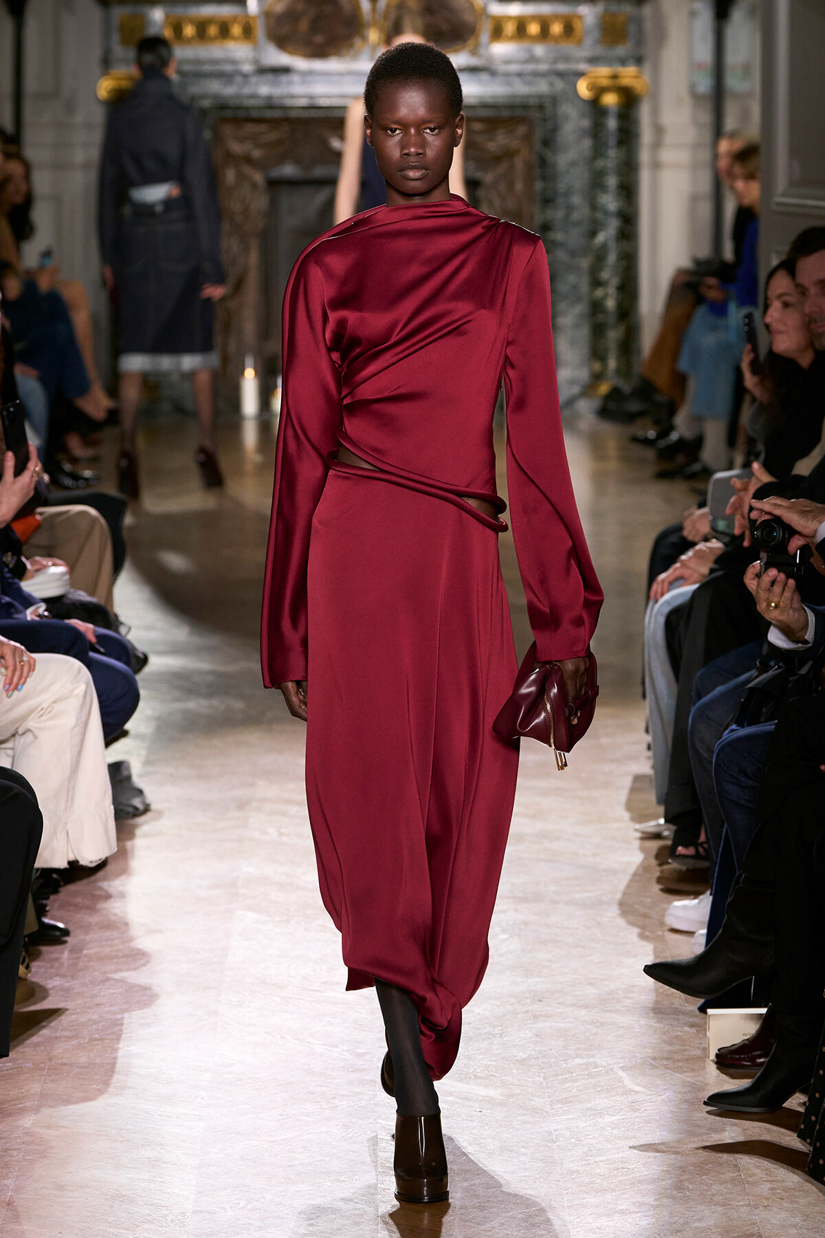 Model walks the runway in a burgundy satin dress with a draped wrap, carrying a small burgundy clutch. Audience lines the catwalk behind.