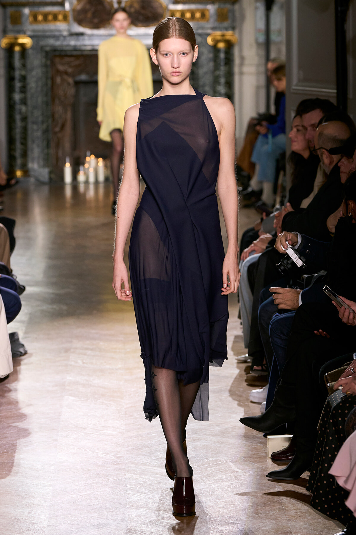 Model walks the runway in a navy sheer dress with geometric panels, audience seated along the catwalk.