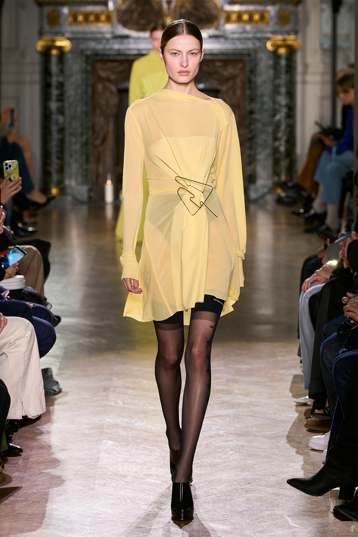 Runway model walks in a pale yellow sheer wrap dress with a sculptural black belt, black tights, and black ankle boots.