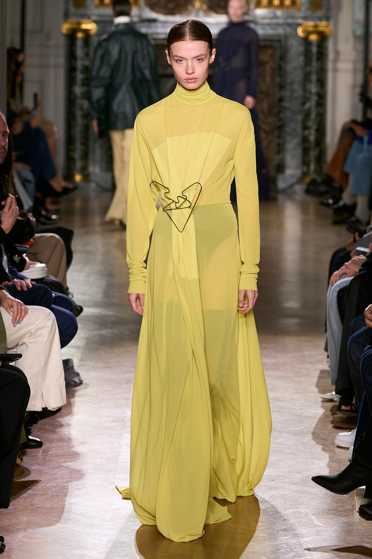 Model on a fashion runway wearing a translucent yellow, high-neck gown with a dramatic, flowing skirt and a geometric metal belt at the waist.