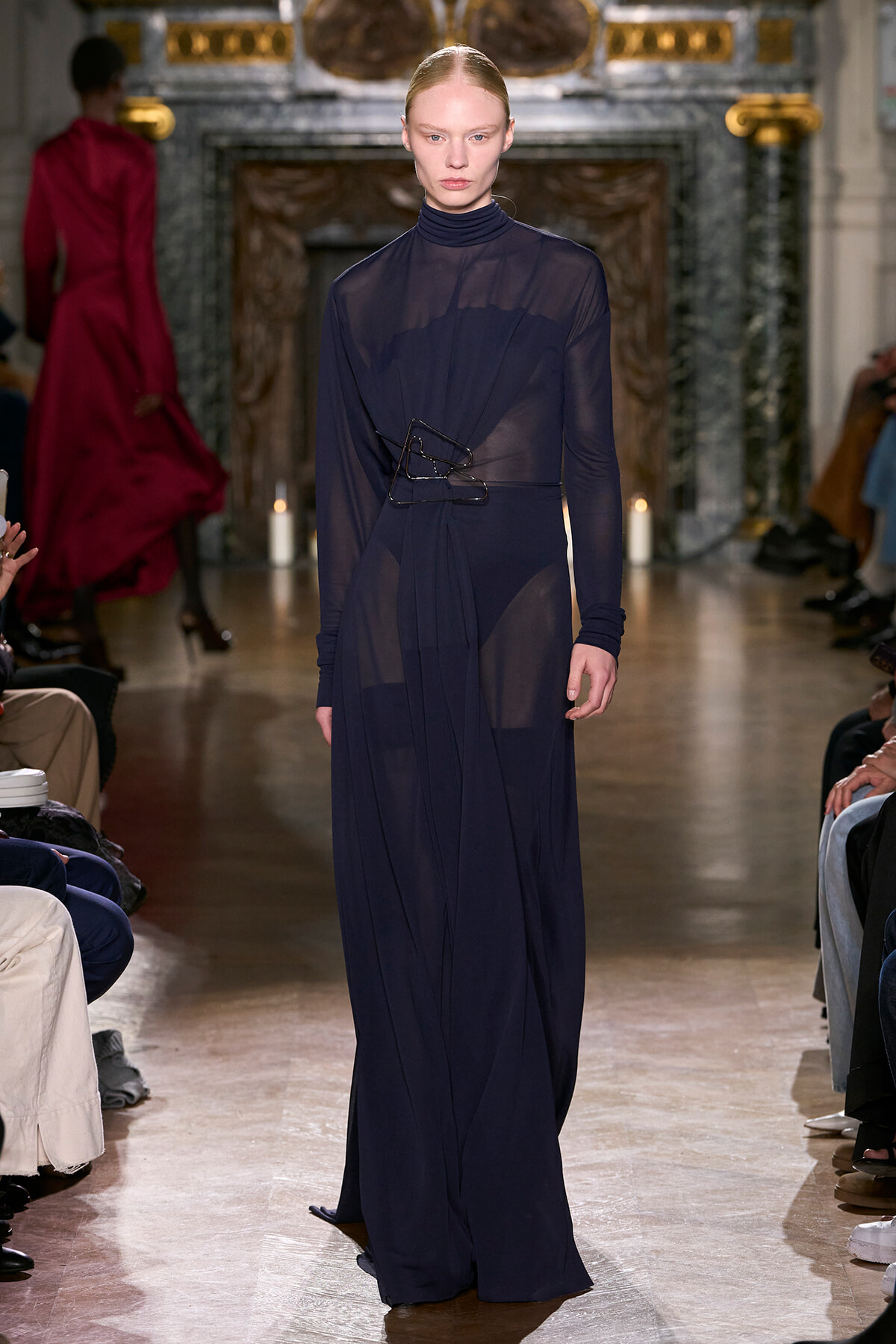 Model walks a dark navy sheer gown with a high neck belt, on a runway in a grand ornate venue.