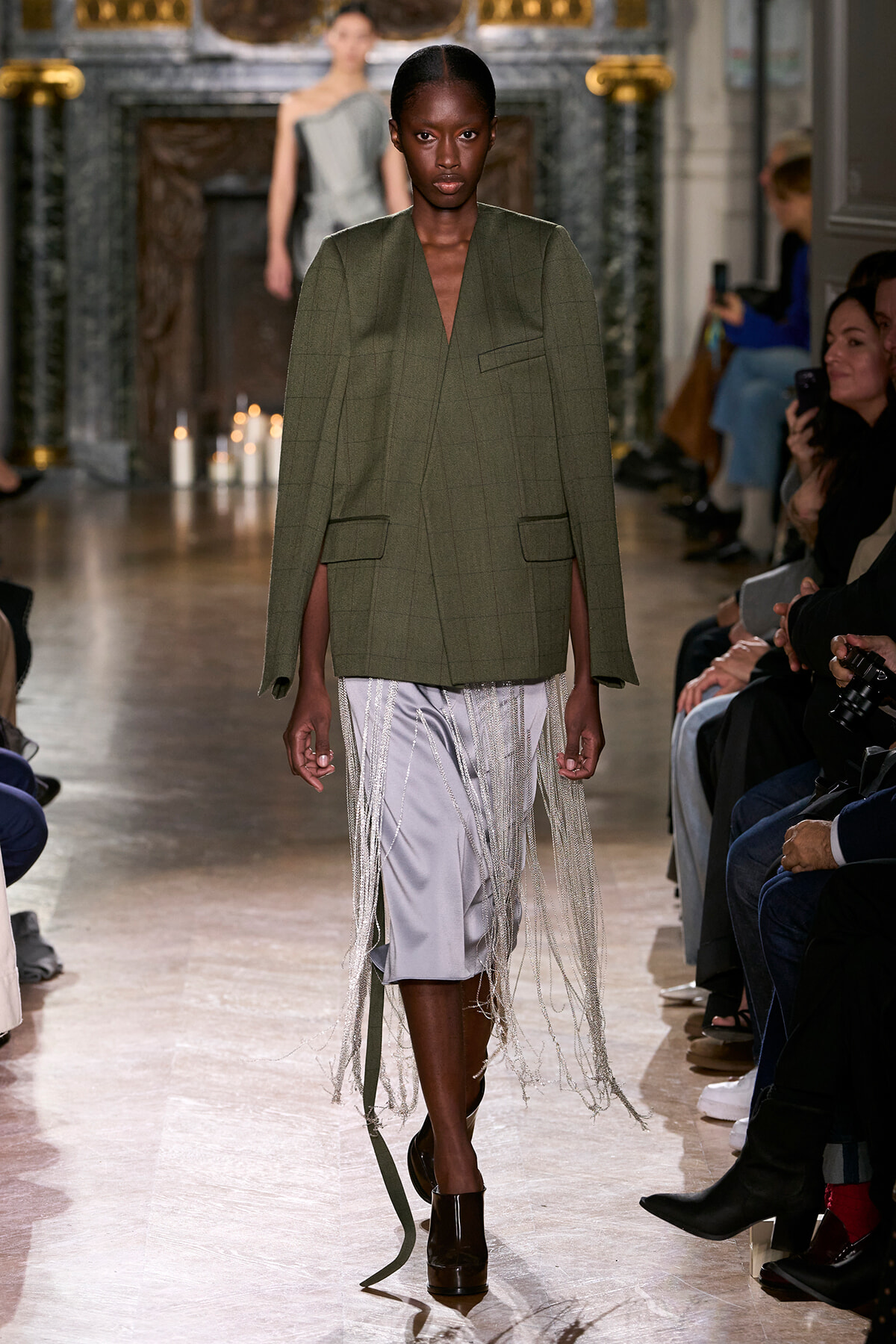 Model walks the runway in an olive green oversized blazer with a long metallic fringe skirt, audience along the sides watch attentively.