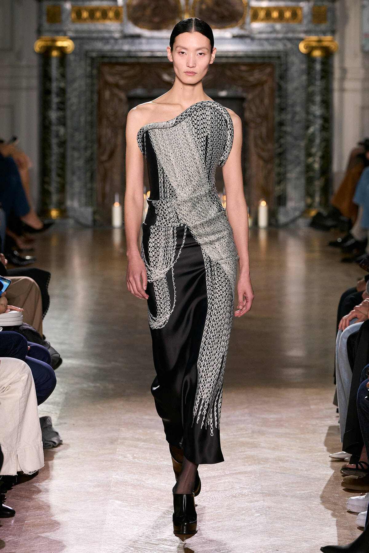 Female model walking a runway wearing a black satin gown with a draped silver chain overlay forming wave-like panels.