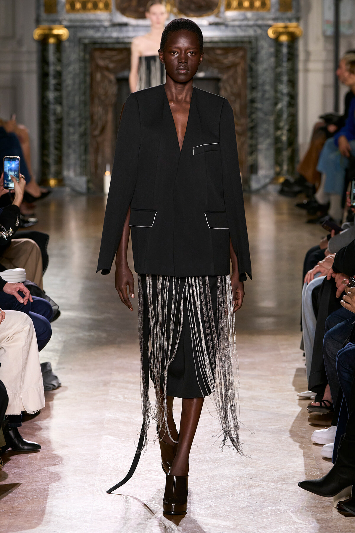 Model walks the runway in a tailored black blazer with white piping and long silver fringe cascading from the waist.