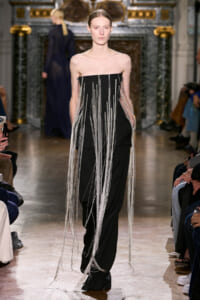 Model on a fashion runway wearing a strapless black outfit with long metallic fringe strands cascading down the front and sides.