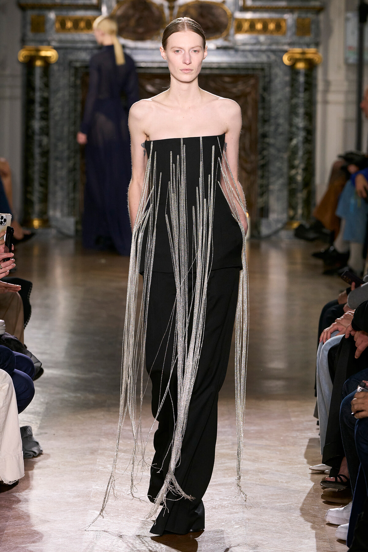 Model on a fashion runway wearing a strapless black outfit with long metallic fringe strands cascading down the front and sides.