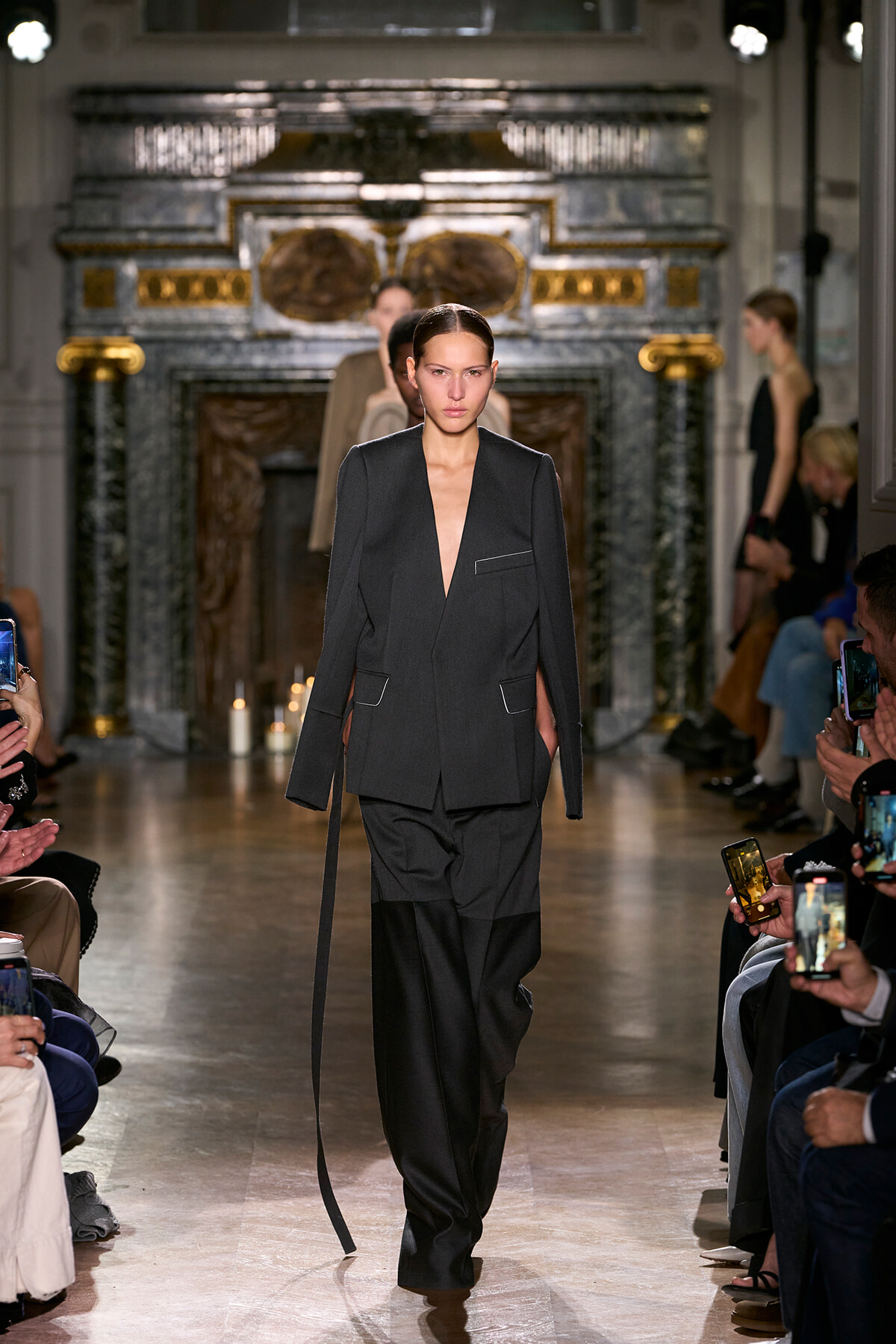 Model walks the runway in a dark tailored blazer and wide-leg pants, hair slicked back, ornate backdrop behind.
