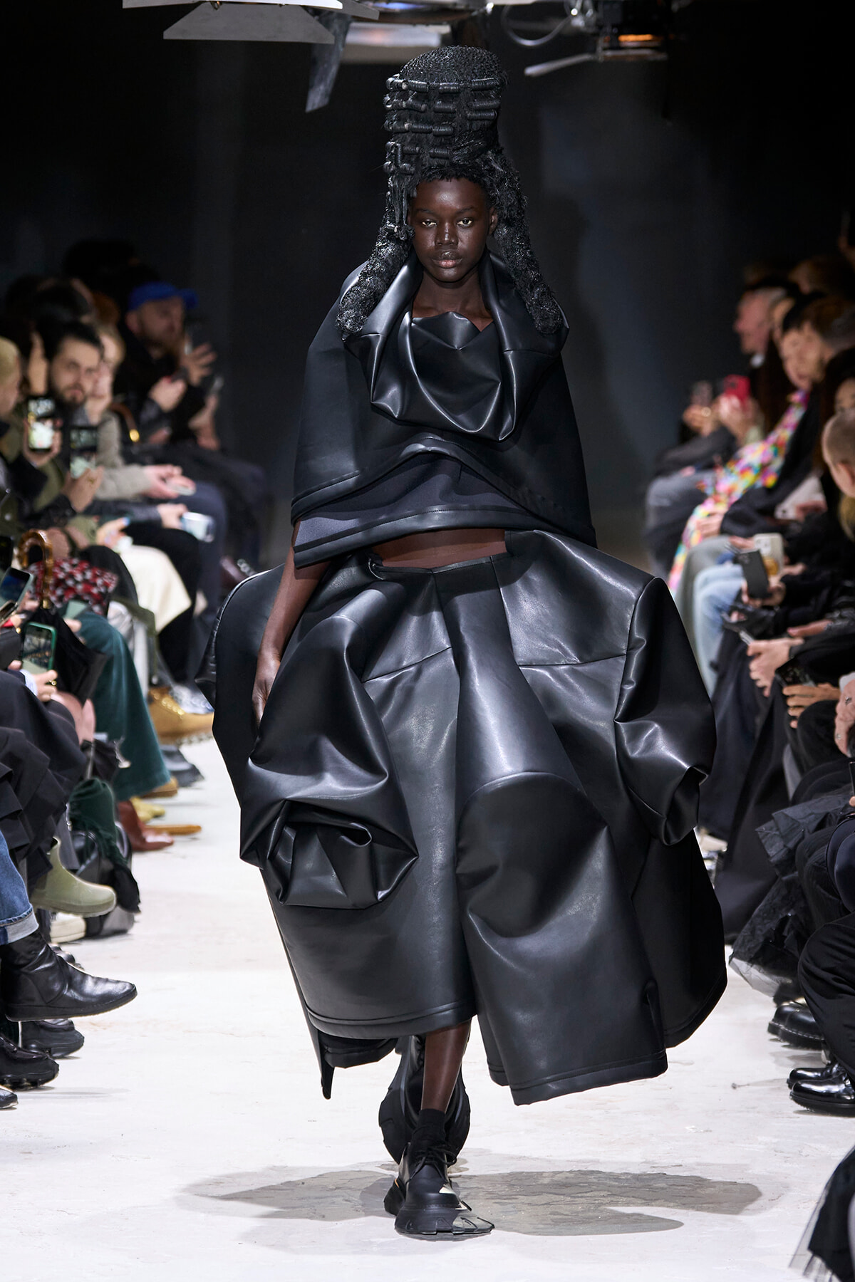 Model walks the runway in an all-black avant-garde leather ensemble with a voluminous sculptural skirt and structured top; audience watches from sides.