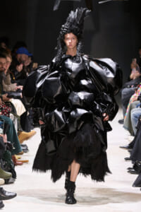 Model on a runway wearing an oversized glossy black gown with sculpted, voluminous sleeves and a tall textured headpiece; audience visible on sides.