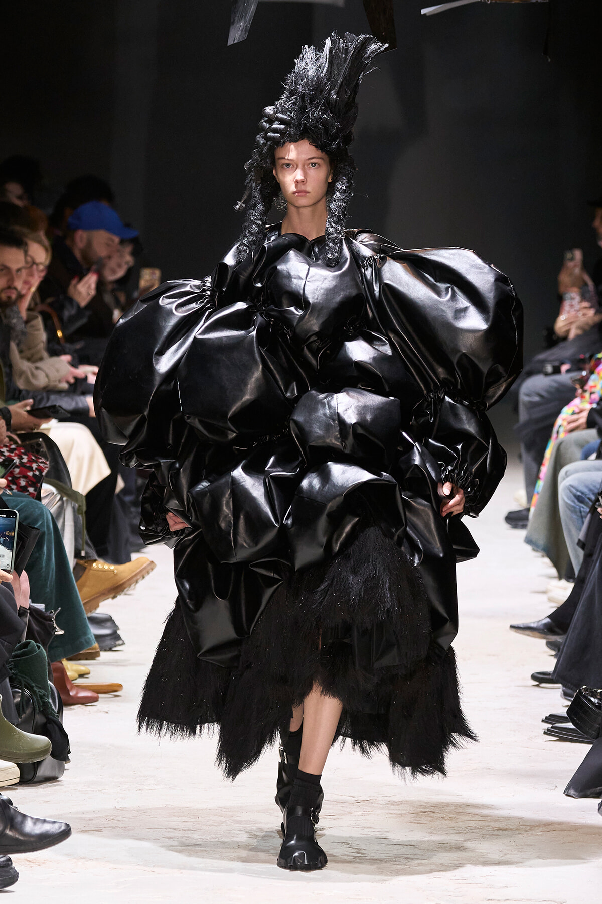 Model on a runway wearing an oversized glossy black gown with sculpted, voluminous sleeves and a tall textured headpiece; audience visible on sides.