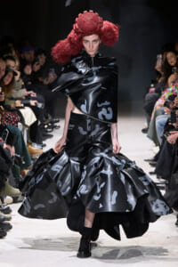 Model walks a fashion runway in a glossy black asymmetric outfit with a voluminous patterned skirt and a sculptural red headpiece, surrounded by spectators.