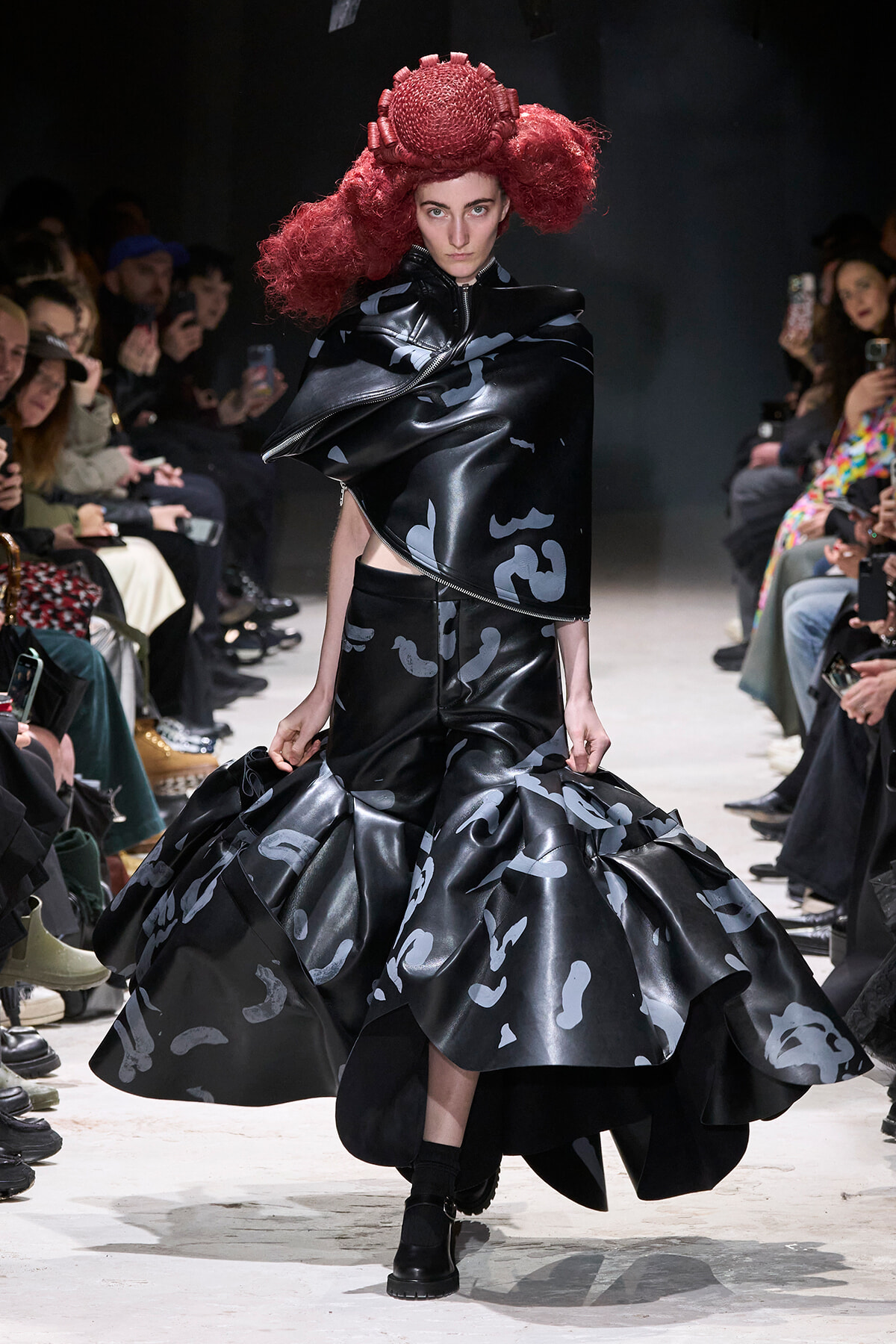 Model walks a fashion runway in a glossy black asymmetric outfit with a voluminous patterned skirt and a sculptural red headpiece, surrounded by spectators.