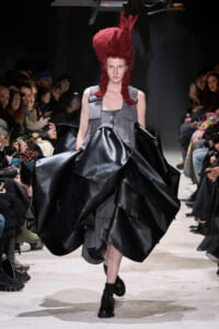 Model walking a runway in a gray patchwork dress with oversized black satin panels and a tall red hair sculpture, flanked by seated audience members.