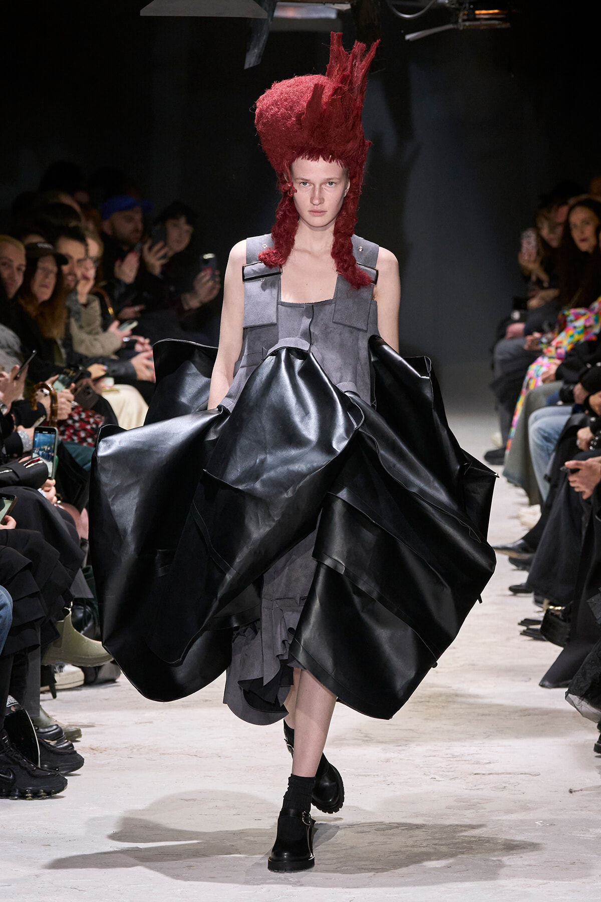 Model walking a runway in a gray patchwork dress with oversized black satin panels and a tall red hair sculpture, flanked by seated audience members.