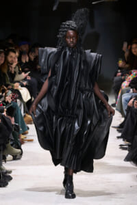 Model walks a fashion runway in a dramatic, glossy black sculptural gown with exaggerated shoulders and voluminous draped folds; audience seated along the catwalk watches.