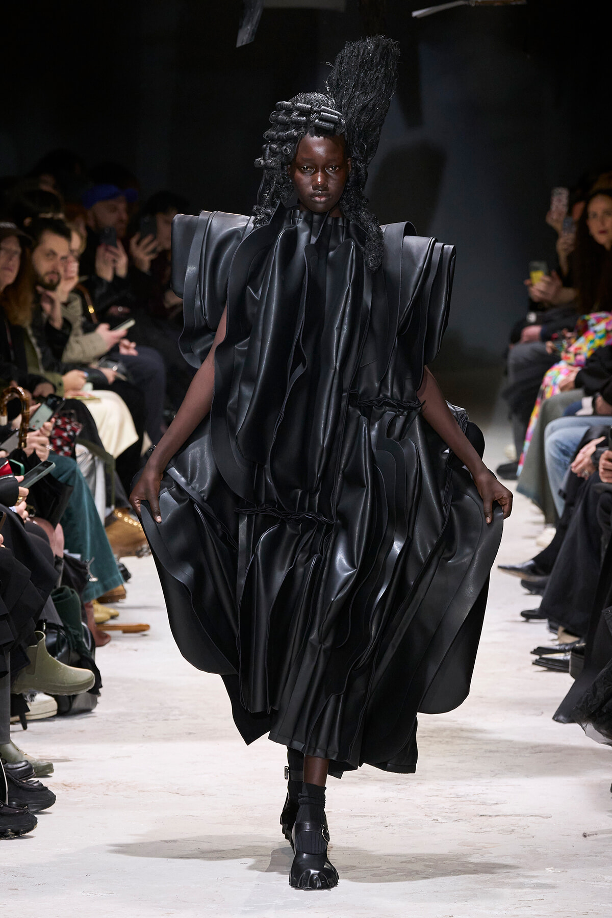 Model walks a fashion runway in a dramatic, glossy black sculptural gown with exaggerated shoulders and voluminous draped folds; audience seated along the catwalk watches.