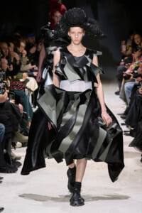 Model walks the runway in a black-gray sculptural dress with oversized layered ruffles and a textured black headpiece on a dark backdrop.
