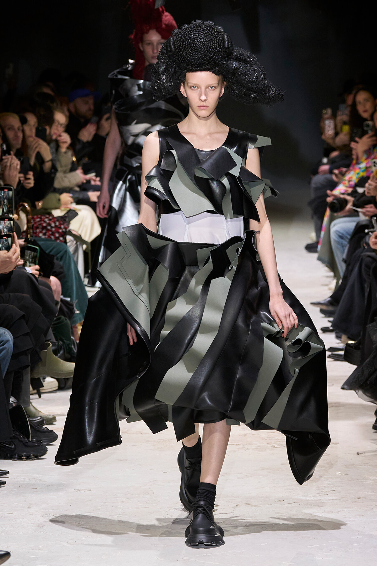 Model walks the runway in a black-gray sculptural dress with oversized layered ruffles and a textured black headpiece on a dark backdrop.