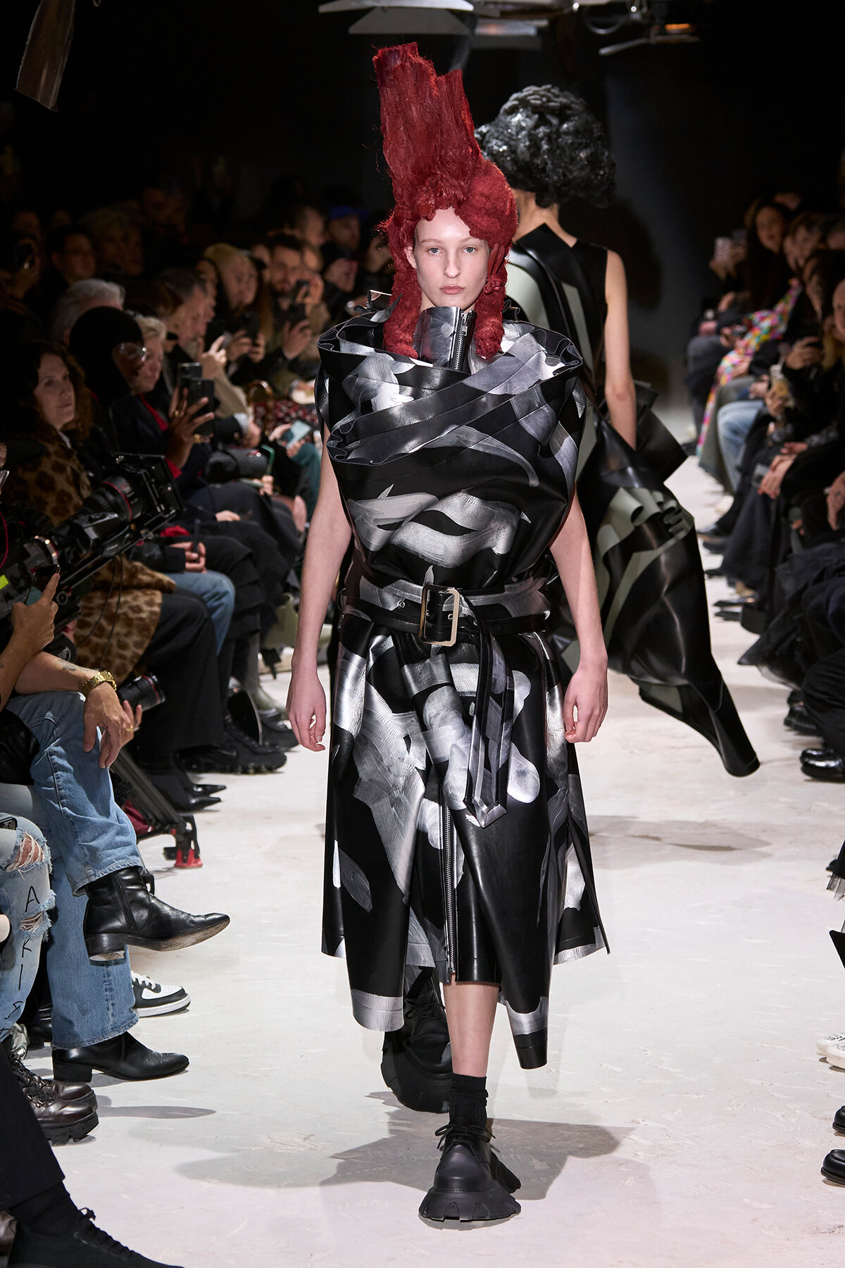 Model walks the runway in a metallic black-silver wrapped dress with a thick belt and dramatic red sculptural hair, audience along the catwalk watching.