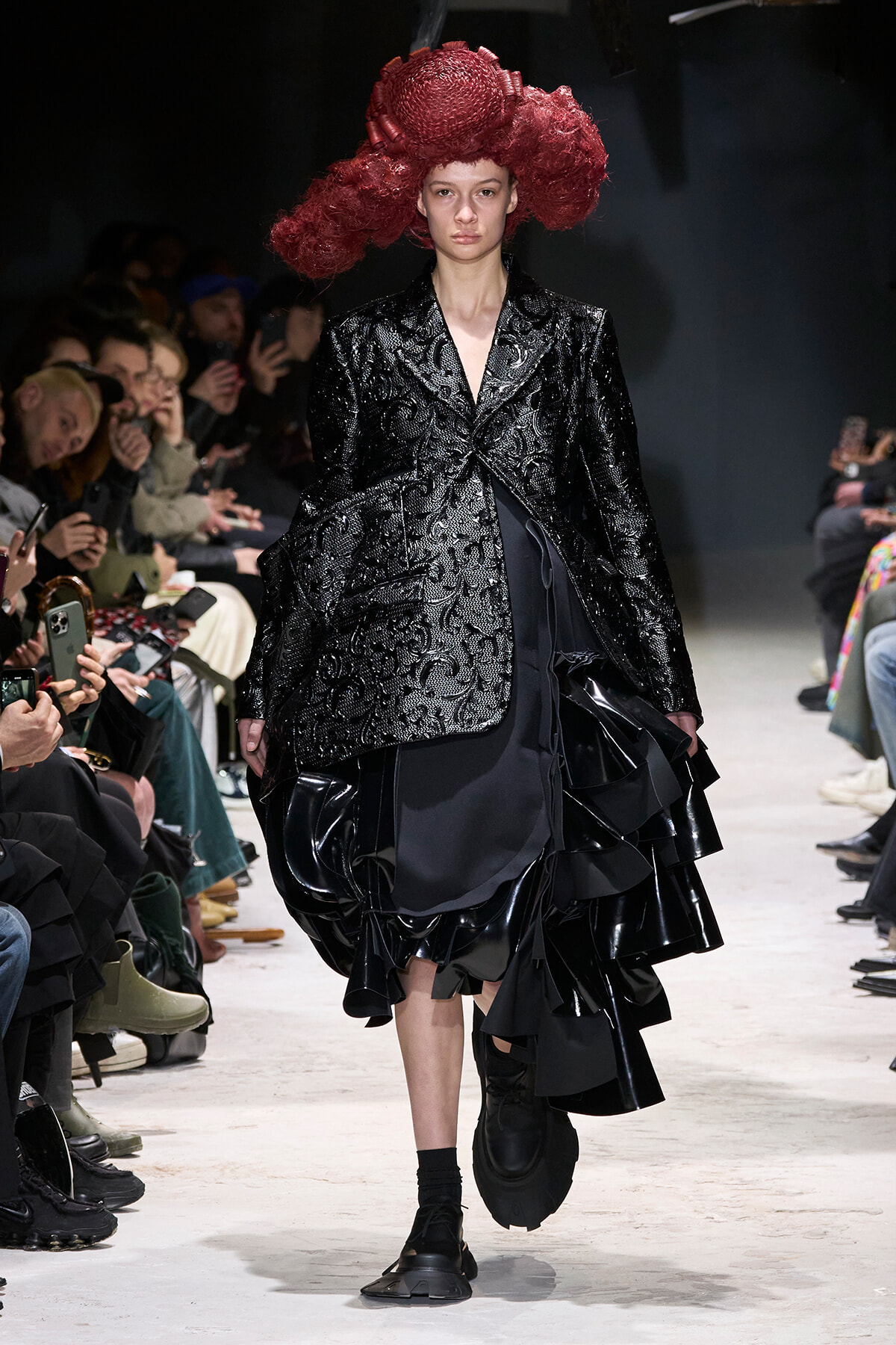Model walks the runway in a glossy black textured blazer with a voluminous red sculptural headpiece; audience photographers line the catwalk.