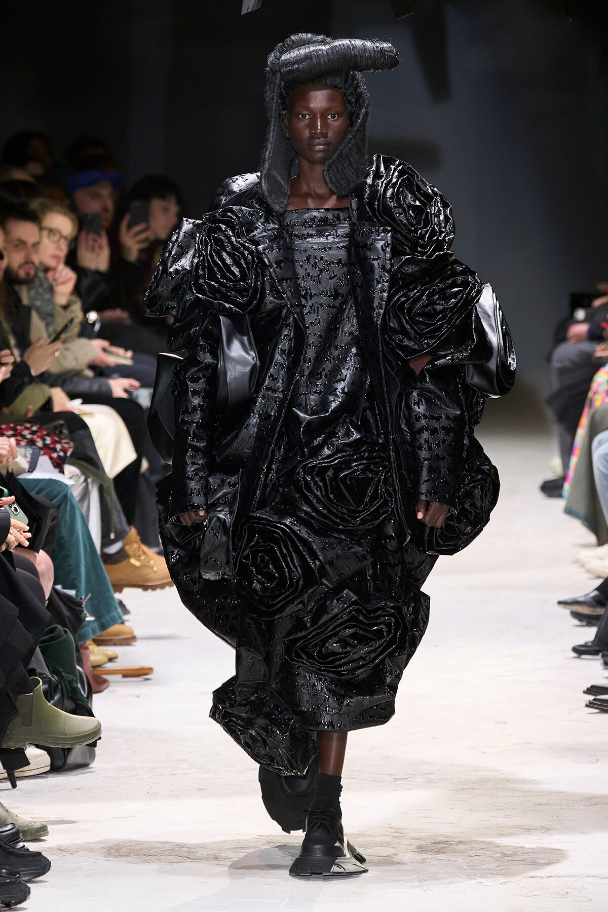 Model walks the runway in a glossy black couture gown with large rosette textures and sculpted hair; audience in the background.