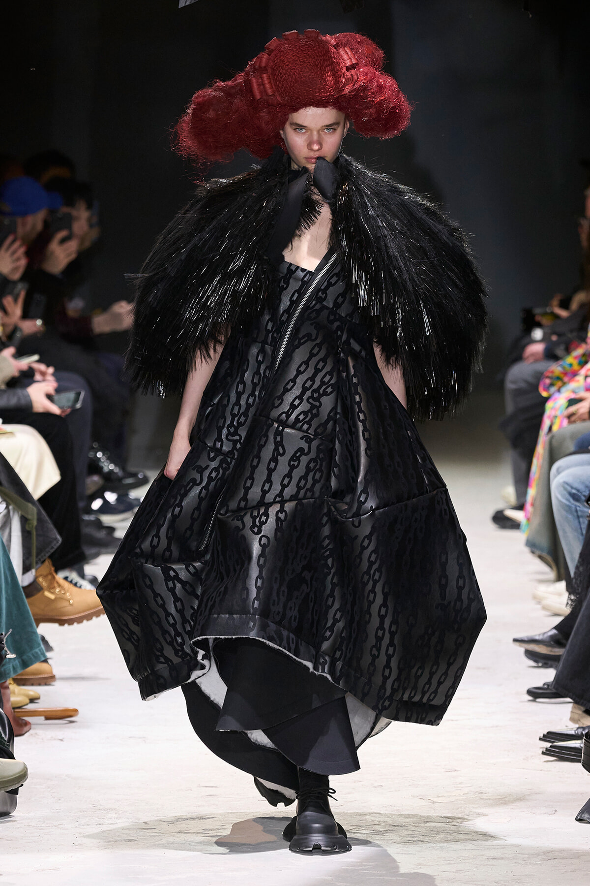 Model walks the runway in a voluminous black patterned dress with a dramatic glossy cape and oversized black fringe, complemented by a red sculptural hat.