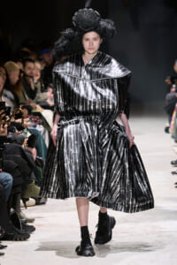 Runway model in a shiny black metallic dress with white barbed-wire pattern and a large sculptural headpiece, walking the catwalk amid seated audience.