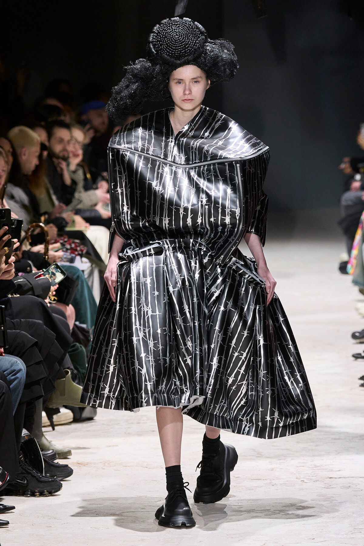 Runway model in a shiny black metallic dress with white barbed-wire pattern and a large sculptural headpiece, walking the catwalk amid seated audience.