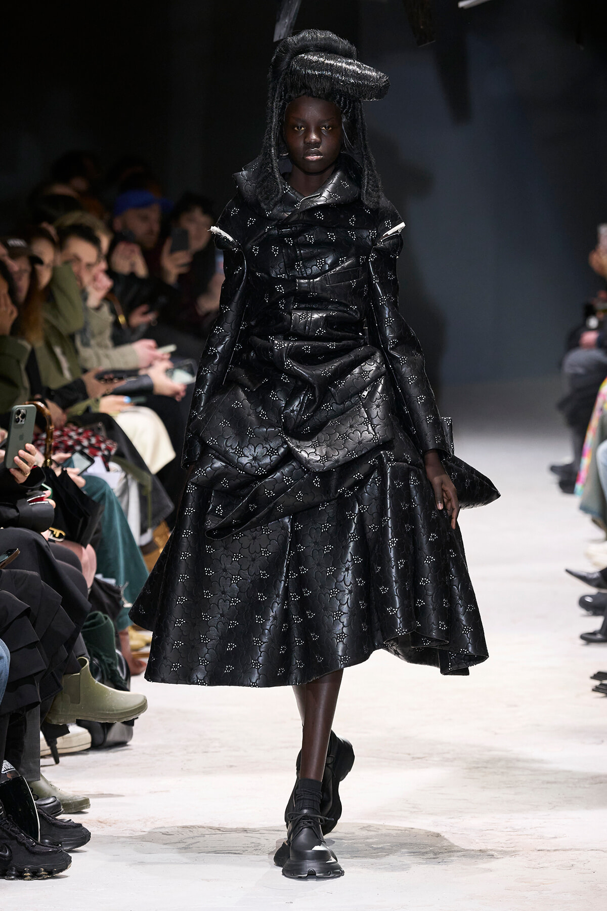 Model walks a fashion-show runway in a voluminous black quilted dress with a floral pattern and a sculptural black headpiece; audience watches from the sides.