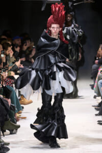 Model walks the runway in a black, glossy, voluminous dress with oversized ruffles and a tall red mohawk, while audience members photograph the show.