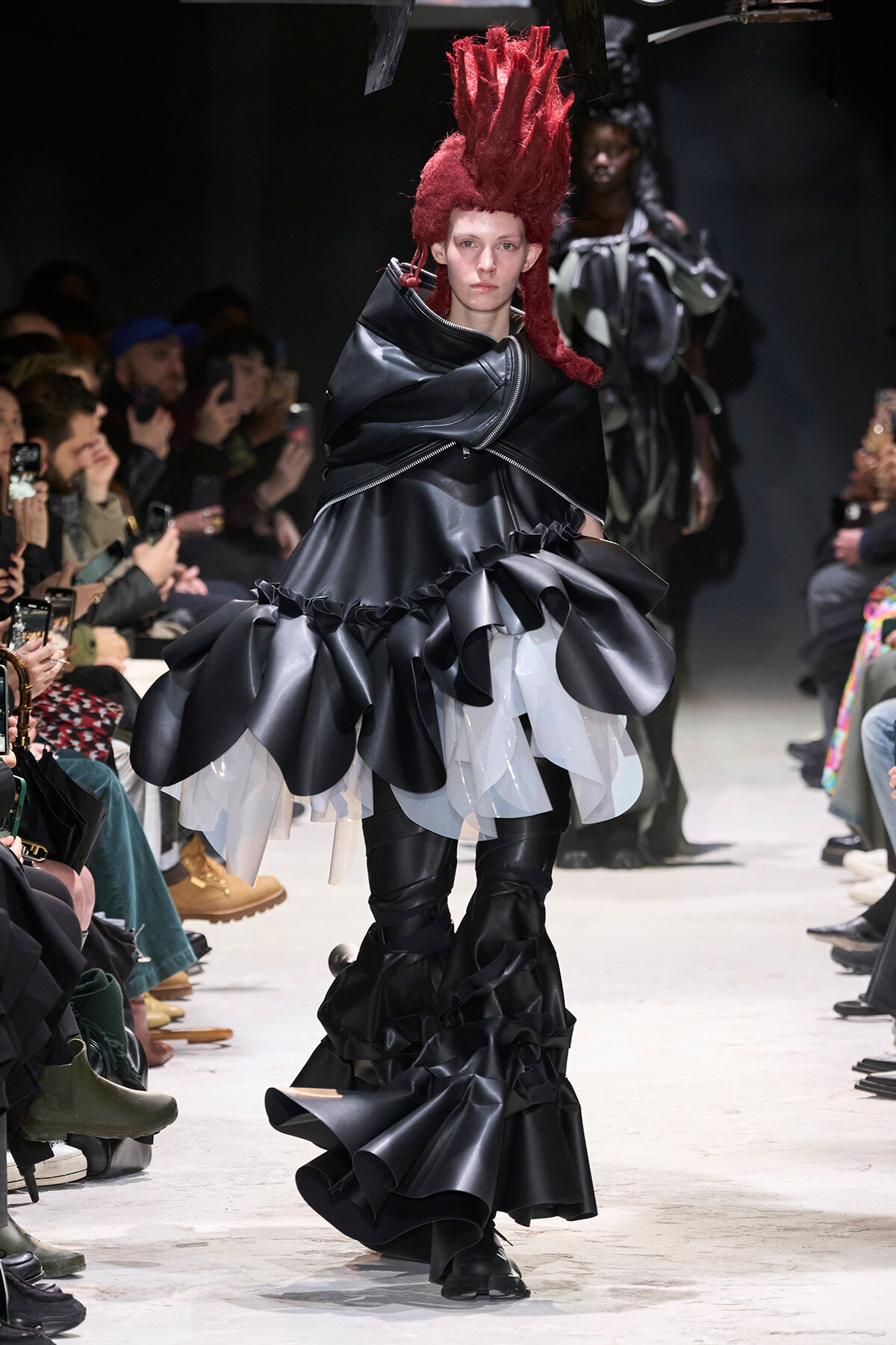 Model walks the runway in a black, glossy, voluminous dress with oversized ruffles and a tall red mohawk, while audience members photograph the show.