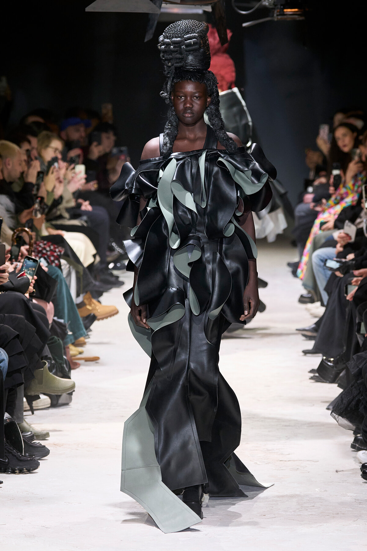 Model walks the runway in a black and sage-green sculptural gown with oversized, layered ruffles; spectators line the catwalk, many taking photos.