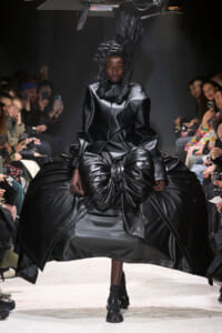 Model on a runway wearing a dramatic black satin dress with a voluminous bow-like skirt and sculpted hair; audience members lean in with phones nearby.