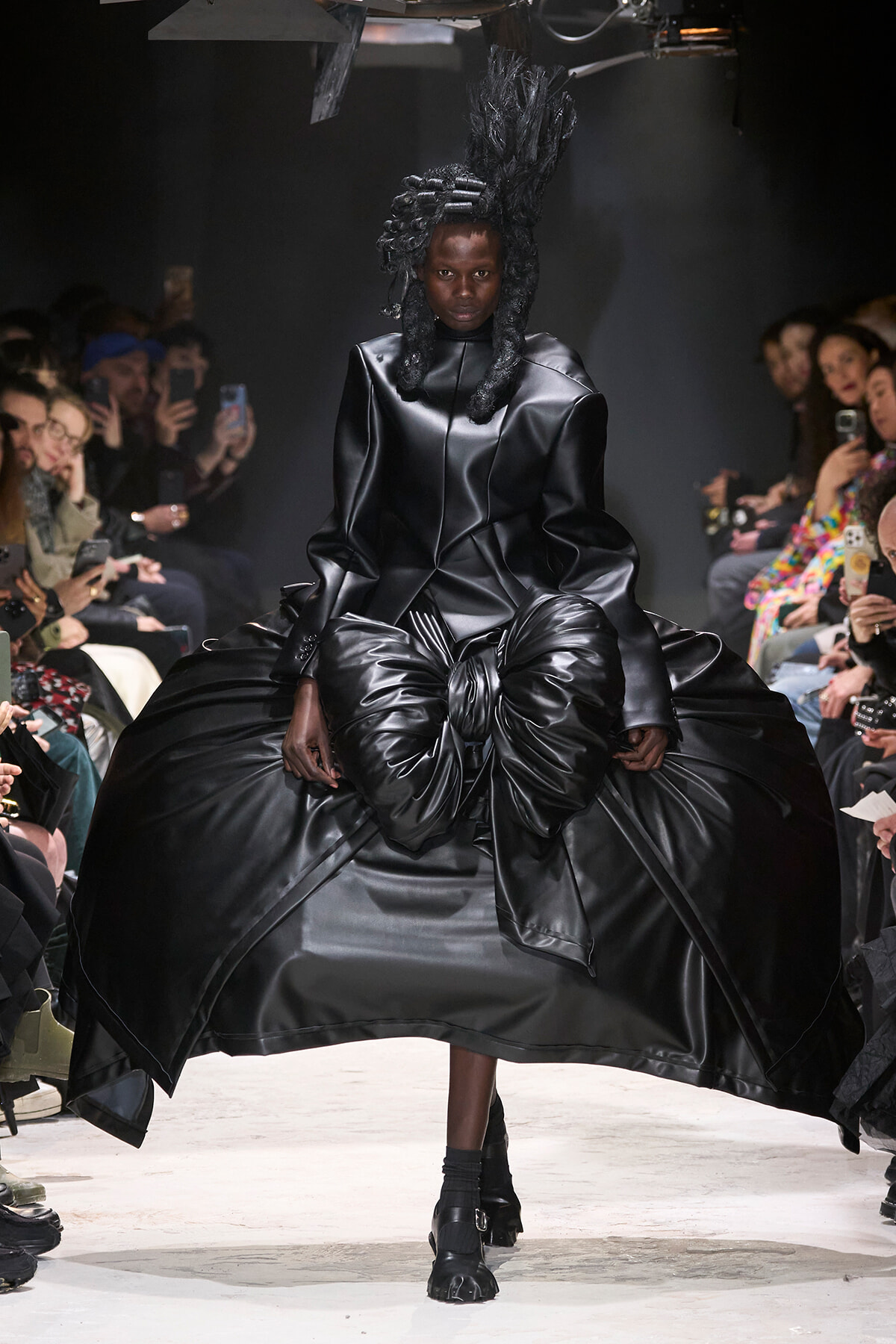 Model on a runway wearing a dramatic black satin dress with a voluminous bow-like skirt and sculpted hair; audience members lean in with phones nearby.