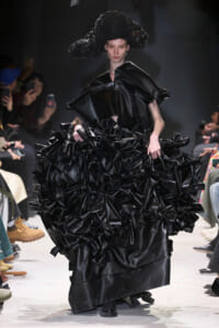 Model on a runway wearing a black glossy gown with a huge, ruffled skirt and a sculptural headpiece; photographers and audience in the background.