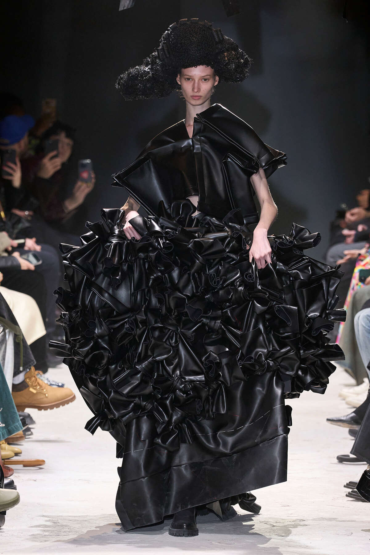 Model on a runway wearing a black glossy gown with a huge, ruffled skirt and a sculptural headpiece; photographers and audience in the background.