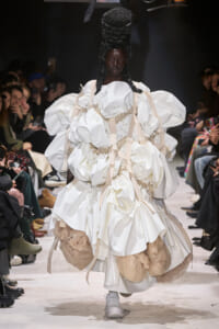 Model on a runway wearing a voluminous white gown with oversized puff sleeves and beige harness-like straps.