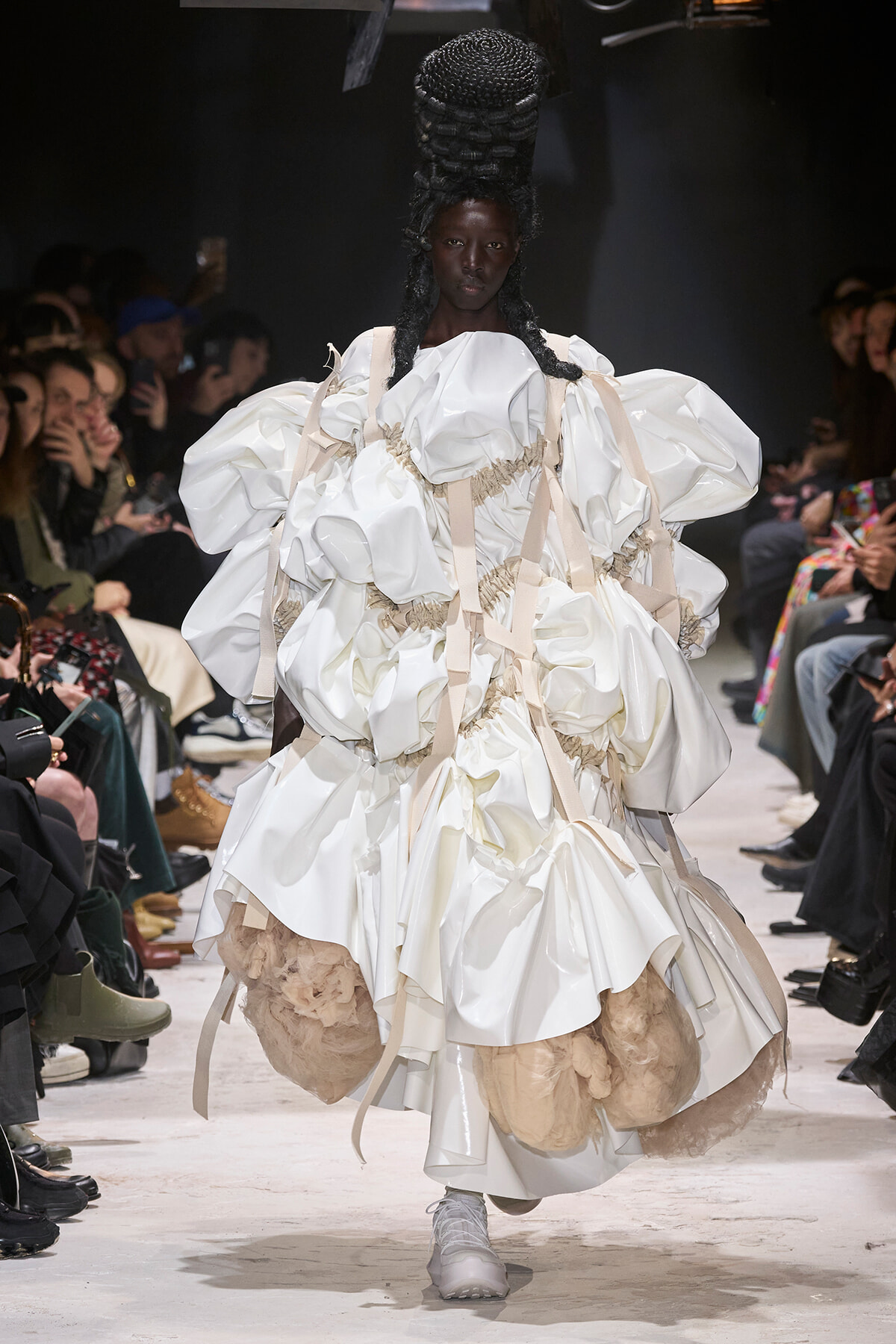 Model on a runway wearing a voluminous white gown with oversized puff sleeves and beige harness-like straps.