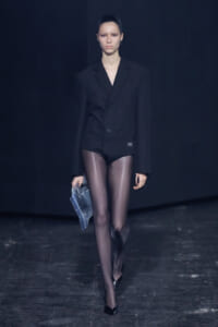 Runway model walking toward the camera in a loose black double-breasted blazer, sheer black tights, black heels, and a blue plastic bag in her left hand.