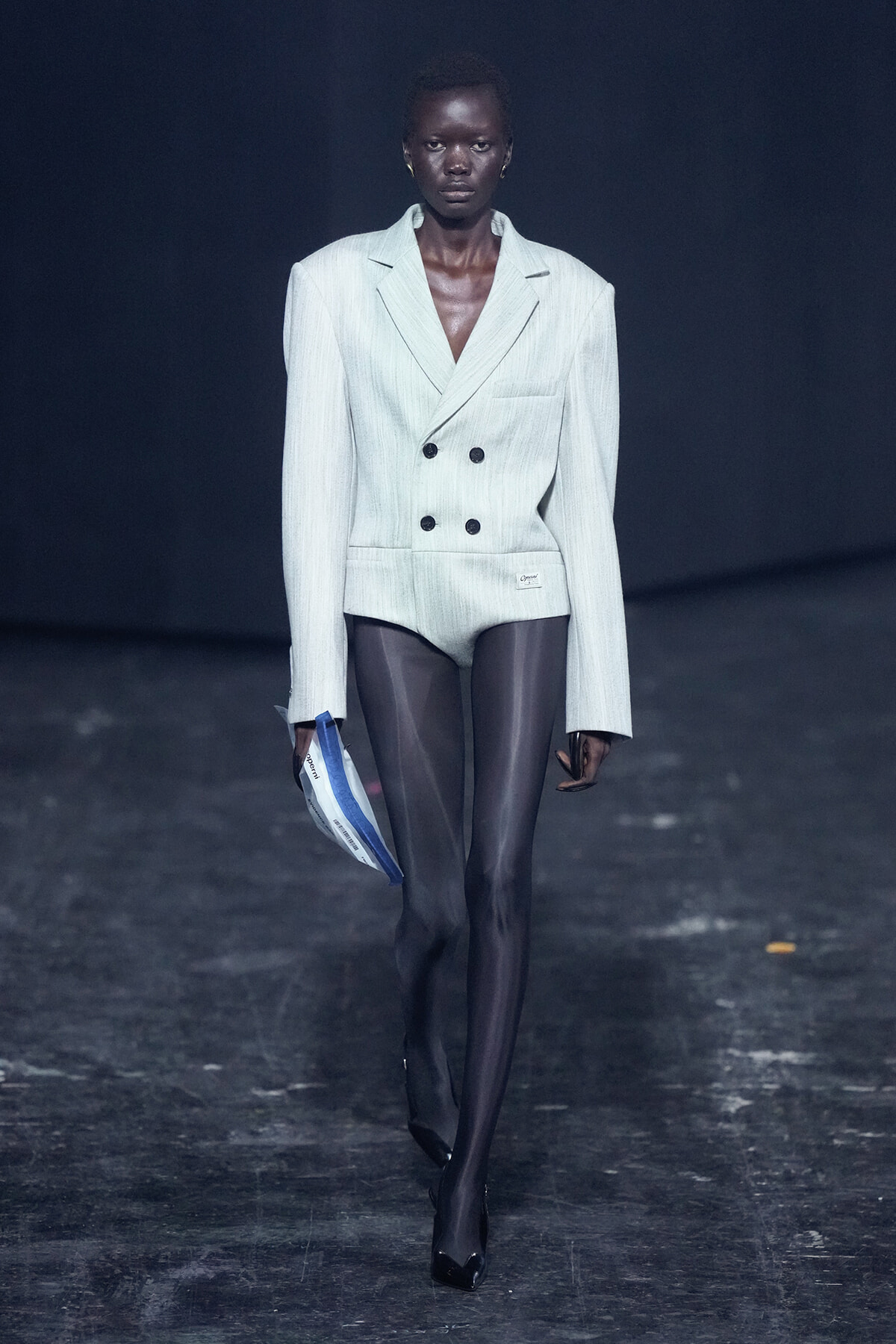 Model walks a runway in a pale pinstripe double-breasted blazer with matching bottoms, sheer black tights, and black heels, holding a blue-and-white program.