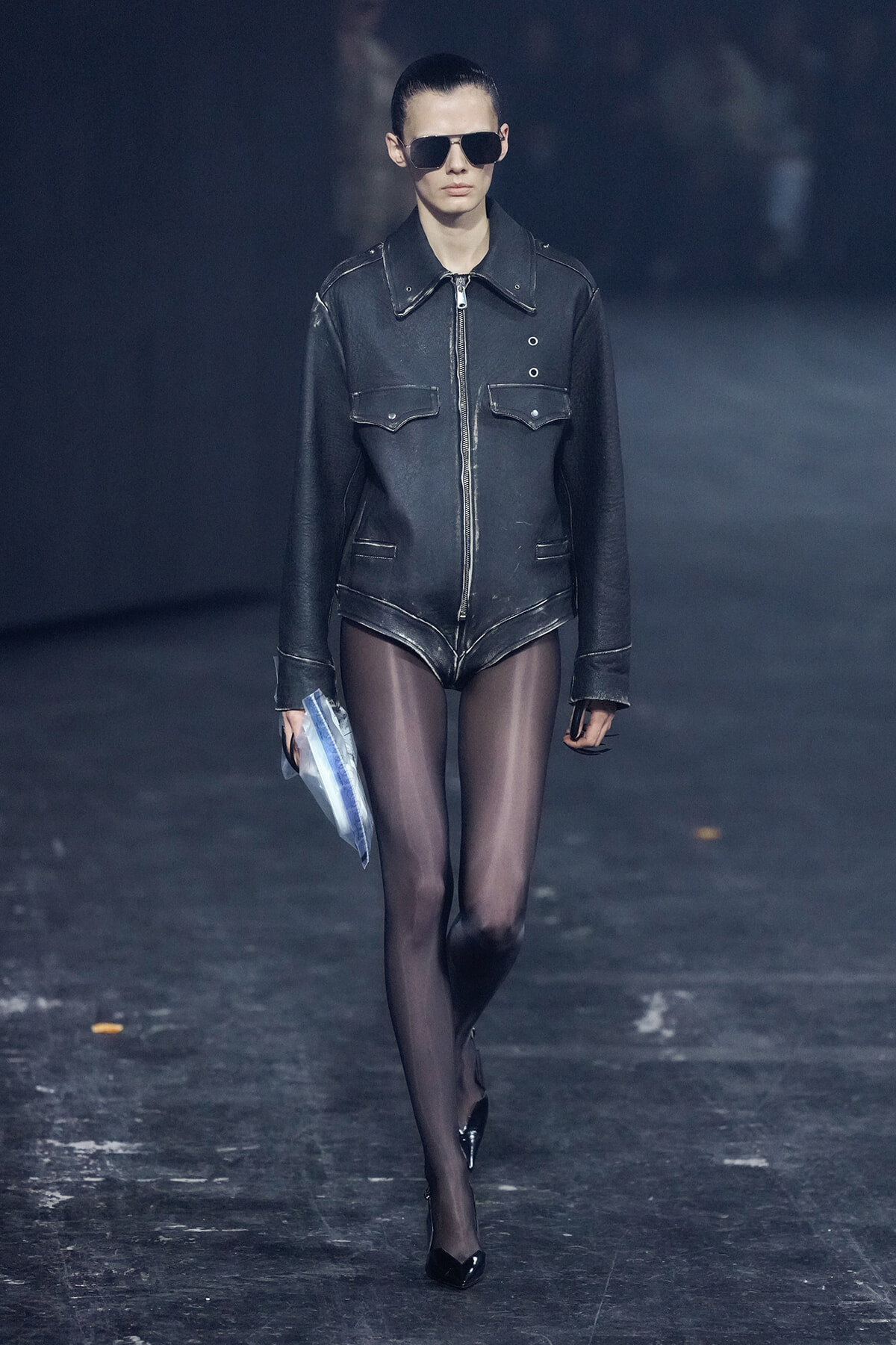 Model walks the runway in a black leather jacket, sheer black tights, black heels, and aviator sunglasses, carrying a blue envelope.