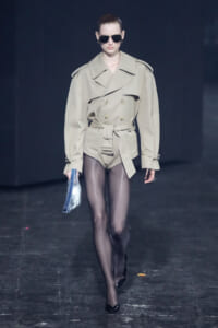 Model walking a fashion runway in a beige trench coat with a belted waist and shoulder epaulettes, wearing black sunglasses and sheer black tights.
