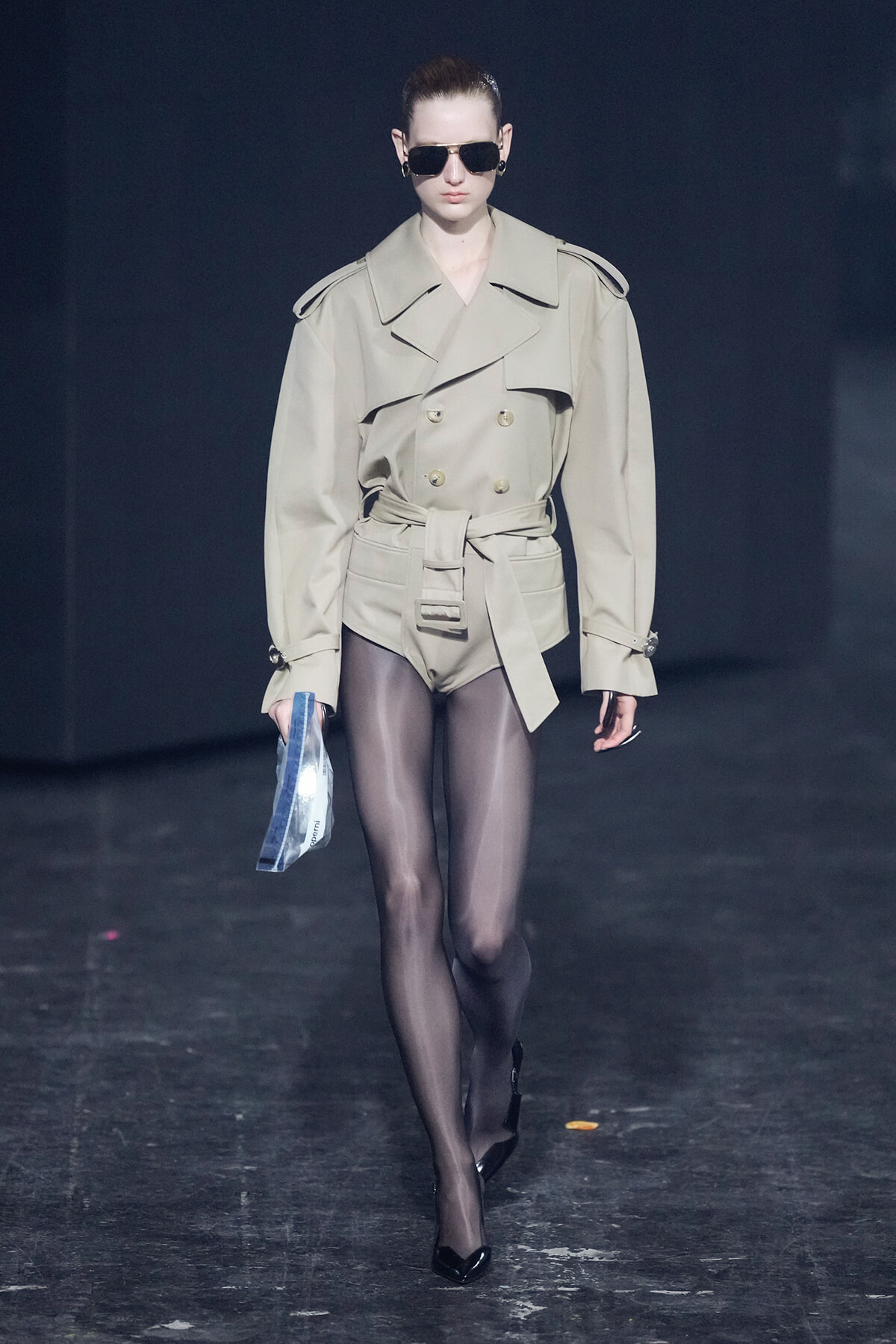 Model walking a fashion runway in a beige trench coat with a belted waist and shoulder epaulettes, wearing black sunglasses and sheer black tights.