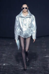 Model walking a runway in a shiny oversized silver jacket, black tights, and sunglasses, carrying a light blue clutch.