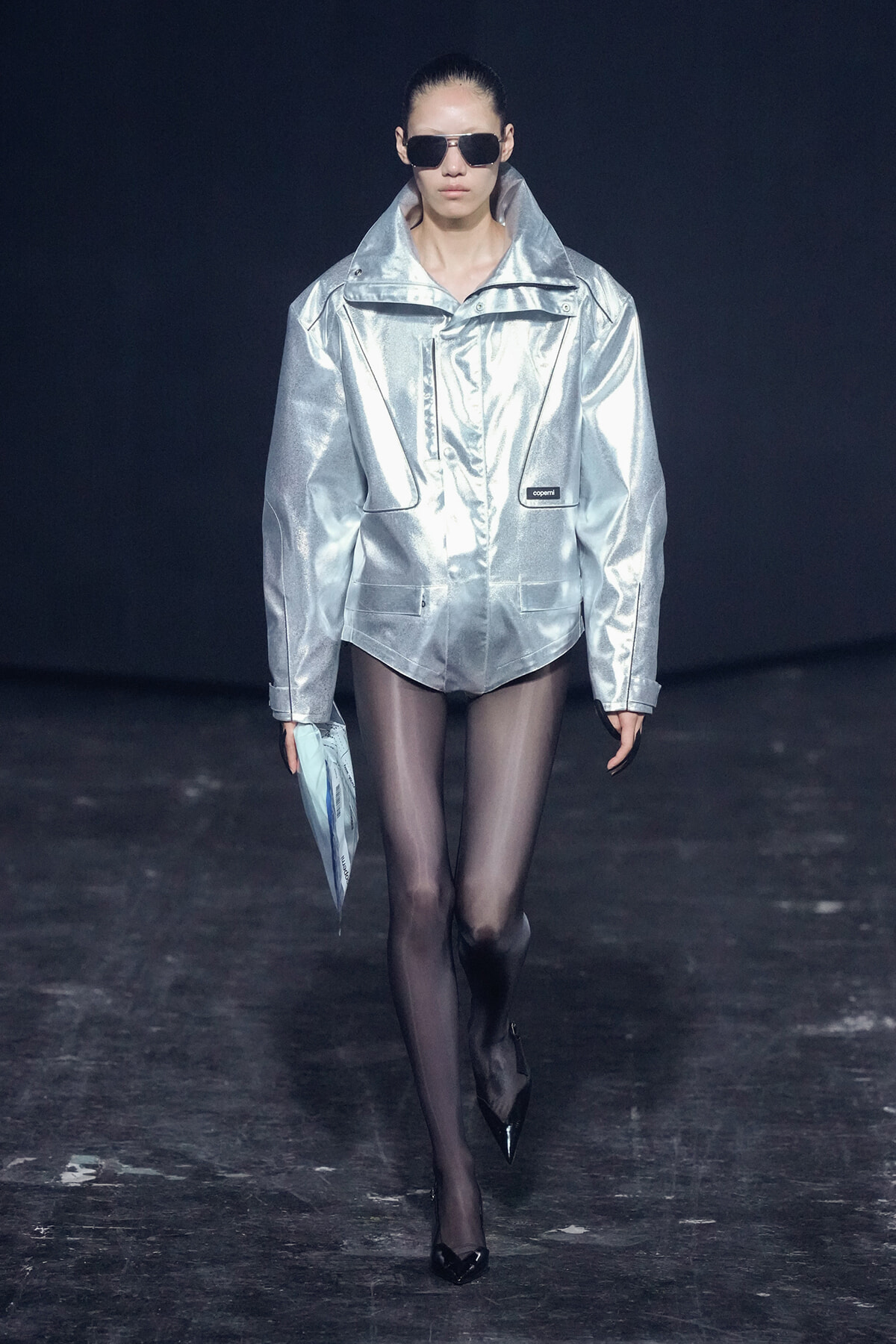 Model walking a runway in a shiny oversized silver jacket, black tights, and sunglasses, carrying a light blue clutch.
