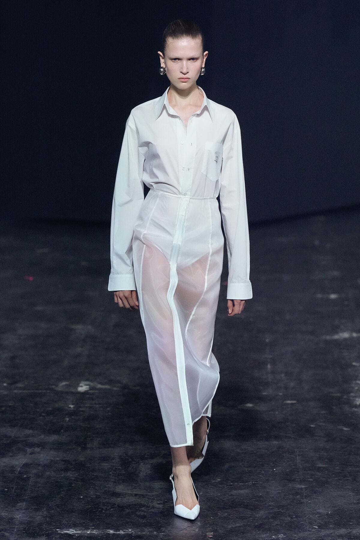 Model walks the runway in a white button-down shirt dress with sheer white pants against a dark backdrop.